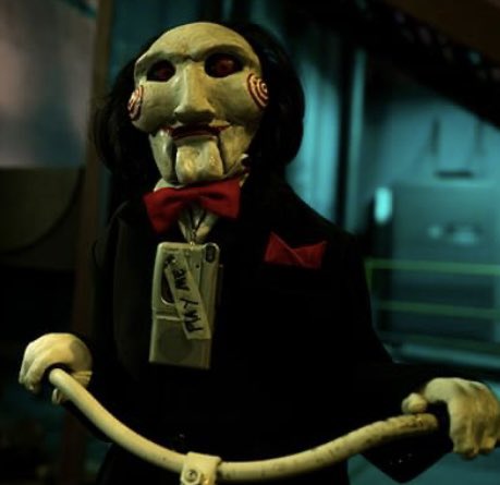 A figure wearing the iconic "Billy the Puppet" mask from the *Saw* horror franchise, dressed in a black suit with a red bow tie, grips the handlebars of a tricycle. A tag reading "Whaddya Hear?" hangs from the mask's neck, evoking the film's cryptic, puppet-driven games—which parallels the post's cryptic message about choosing to believe in a LUNC and USTC altseason, suggesting the fate of the market is left to the viewer's faith.