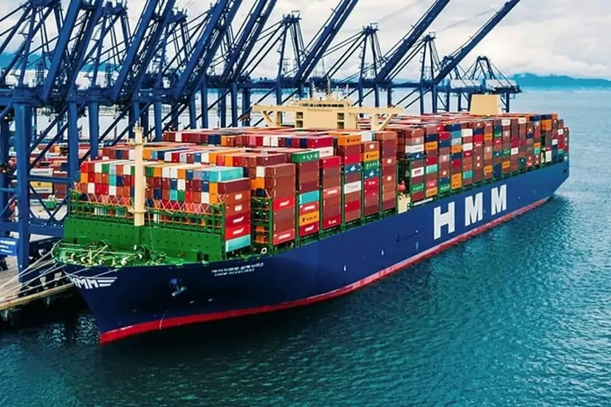 A large HMM container ship loaded with colorful shipping containers is docked at a port with blue cranes towering overhead.