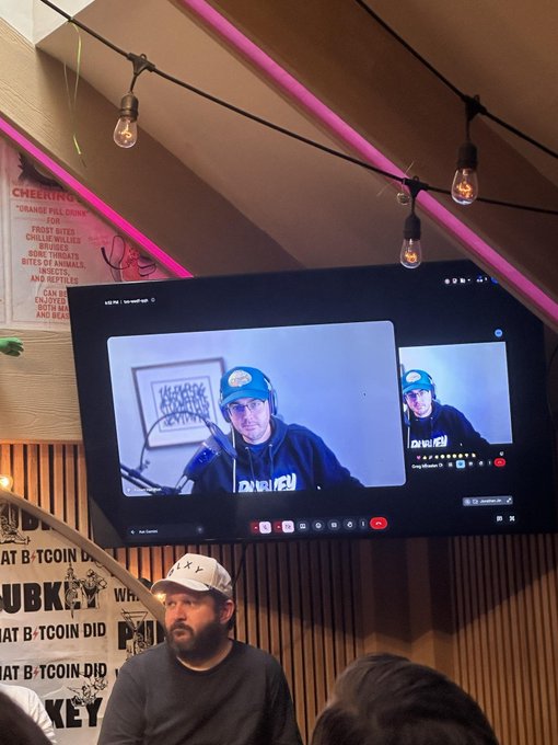 A live podcast recording at PubKey featuring The Blue Matt, intangiblecoins, and darosior in a video call on screen, with an attendee wearing a PubKey hat seated below the display.