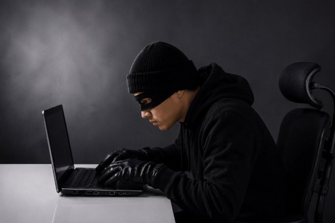 A stereotypical 'hacker' in a black beanie and mask types on a laptop in a dark room, evoking cybercrime tropes.