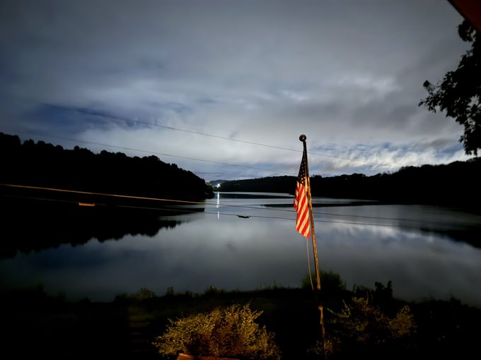 An American flag stands illuminated in front of a calm lake at dusk, with silhouetted hills and a dramatic cloudy sky reflected in the water.