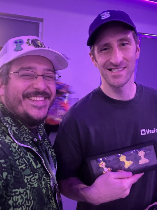 Two smiling men posing together indoors; the one on the right wears a 'Veefr' t-shirt and holds a device displaying pixelated character art, while the man on the left wears a colorful patterned jacket and a cap with stickers.
