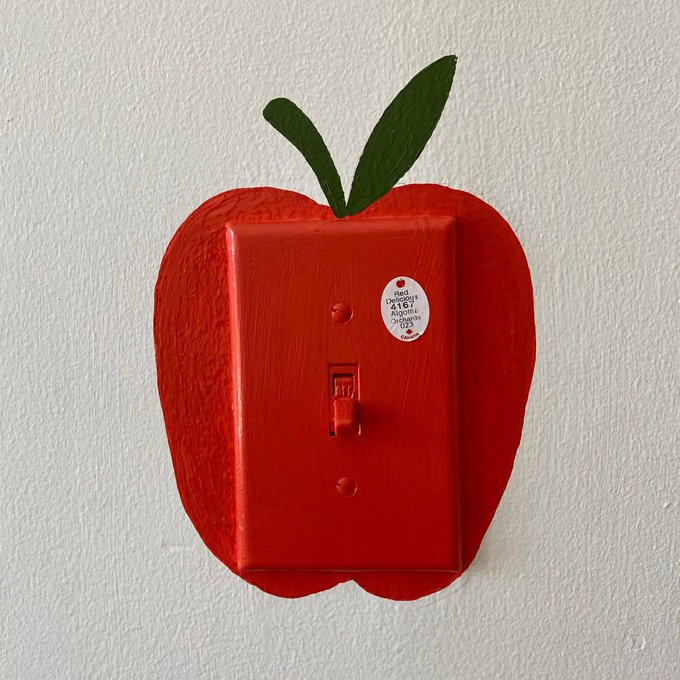 A red light switch plate mounted on a wall, creatively designed to look like an apple with green leaf-shaped extensions at the top and a small 'Red Delicious 0167' sticker on it.