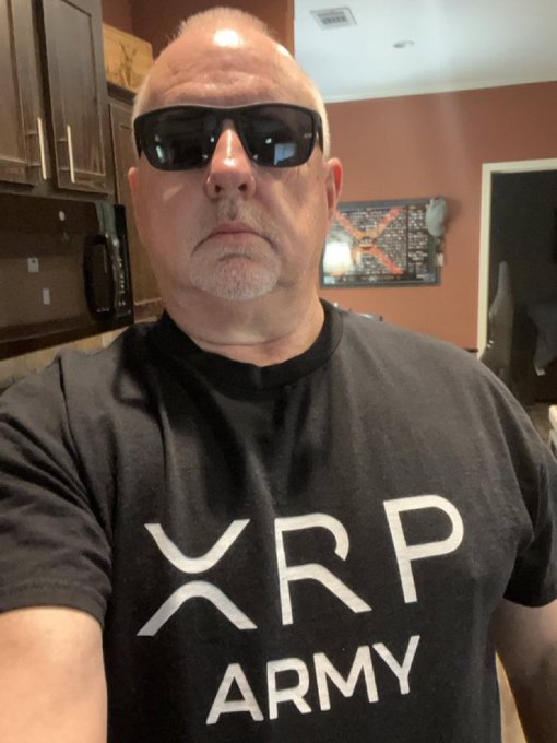 The image features a man wearing a black t-shirt with white text that reads "XRP ARMY!". The man has sunglasses on his head, giving him a cool and confident appearance. He appears to be posing for a photo or taking part in an online discussion about the XRP currency.