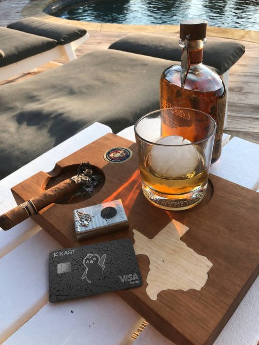 A glass of whiskey with a large ice sphere and bottle of spirits sit on a wooden cigar ashtray tray beside a KAST Visa card and a lighter, with a pool and lounge chairs visible in the background—visually reinforcing the post's message of effortless crypto-powered luxury.