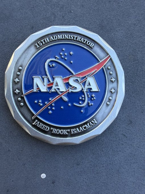 A commemorative NASA challenge coin features the iconic "meatball" logo and is engraved with "15TH ADMINISTRATOR" at the top and "JARED 'ROOK' ISAACMAN" at the bottom, presented by Jared Isaacman to the recipient.