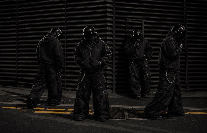 Four figures dressed in black baggy clothing, masks, and helmets with glowing eyes are posed on a street in front of a closed metal shutter.