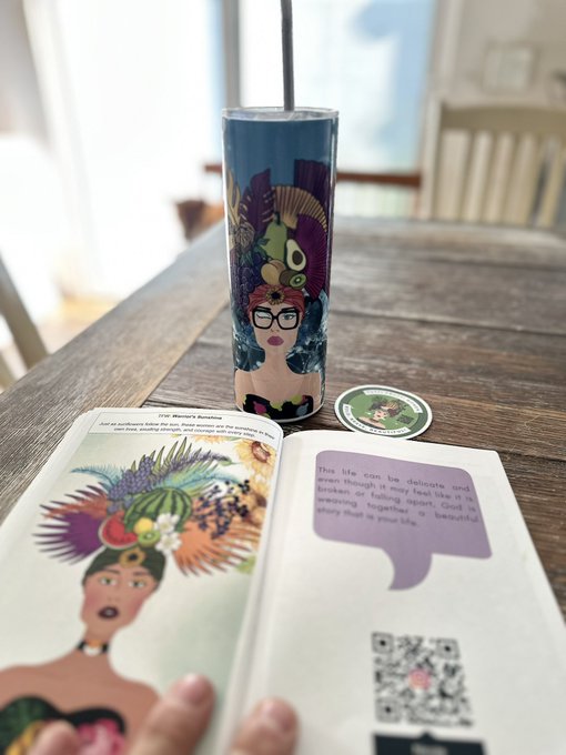 A Frida Kahlo-themed tumbler with a straw sits on a wooden table next to an open art book about Frida, displaying colorful illustrations and a QR code.