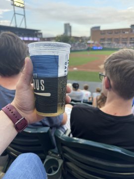 A hand holding a 'Liquid Death' canned beverage at a baseball stadium during a game, with the field and other spectators visible in the background.