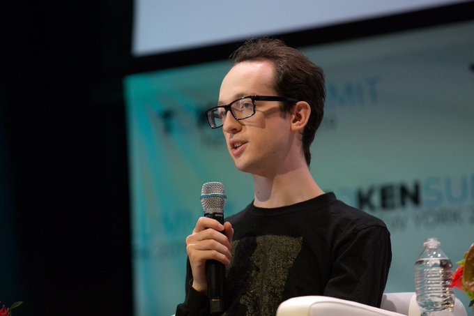 A young man with glasses speaks into a microphone at what appears to be a conference, with partial text "KEN SU" and "SUMMIT" visible on the backdrop behind him. The image relates to the Uniswap context by showing what could be a speaker at a crypto/blockchain summit discussing industry topics like the domain acquisition controversy.