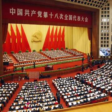 The image shows the 18th National Congress of the Chinese Communist Party, as indicated by the Chinese characters "中国共产党第十八次全国代表大会" displayed prominently on a red banner above the stage, where delegates are seated in a large hall with red flags and a hammer and sickle emblem. The image relates to the post by visually establishing the context of China's central government decision-making, highlighting the authoritative source of the directive to build a national blockchain network as part of the country's economic blueprint.
