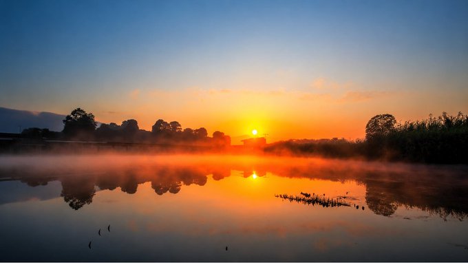The image shows a beautiful sunrise over a lake with foggy water. The sun is rising behind the trees, casting an orange glow across the sky as it sets below the horizon. The scene is captured from a distance, providing a panoramic view of the landscape and the reflection of the sun on the water's surface.
