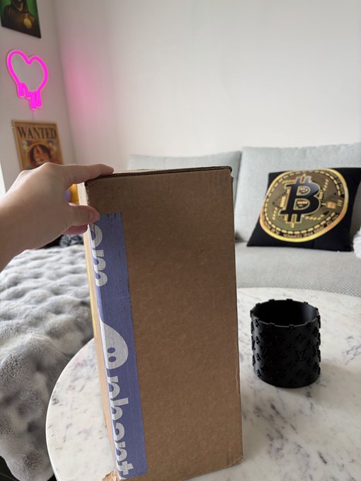 A hand holds a sealed cardboard shipping box with blue and white "about:blank" branded tape on a marble table, with a Bitcoin-themed pillow visible in the background. The setup suggests someone unboxing a delivery from the AboutBlank brand, captioned as if expecting a payout.