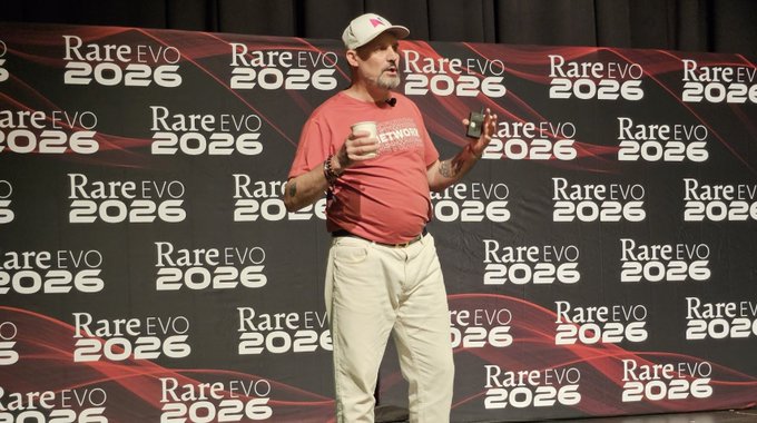 A man wearing a red t-shirt, khaki pants, and a baseball cap speaks on stage at the 'Rare EVO 2026' conference, holding what appears to be a can and a small device.