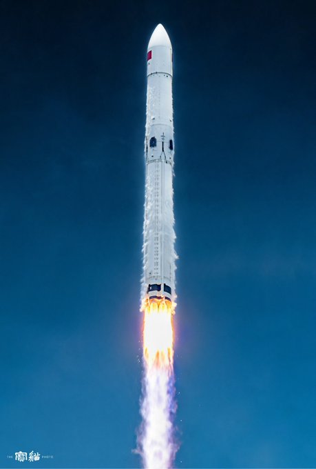 A Chinese Long March 10B (CZ-10B) rocket ascends against a deep blue sky, its engines blazing a brilliant plume of flame as white vapor streams down its fuselage. The image captures the vehicle mid-launch, showcasing the powerful thrust characteristic of China's next-generation crewed lunar launch system.