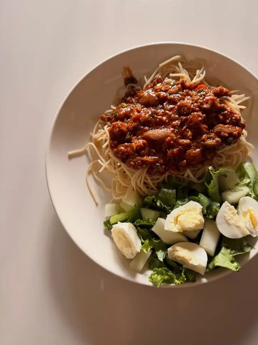A white bowl contains spaghetti topped with a chunky meat sauce, served alongside a side salad of fresh greens and sliced hard-boiled eggs. The comforting, homemade meal suggests a simple "okay" kind of day fueling the creator for tomorrow's hustle.