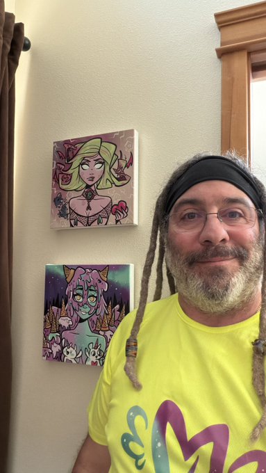 A man with dreadlocks and glasses smiles at the camera, wearing a yellow t-shirt. Behind him on the wall hang two small square paintings featuring stylized female characters with vibrant hair.