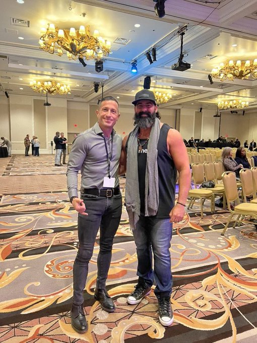 Two men pose together at a conference or convention in a grand ballroom with chandeliers and ornate patterned carpeting; one wears a gray shirt with a lanyard and the other wears a dark tank top with a gray scarf.