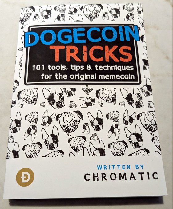 A physical paperback copy of "Dogecoin Tricks: 101 tools, tips & techniques for the original memecoin," written by Chromatic, rests on a marble surface with a cover featuring the iconic Shiba Inu dog pattern—highlighting the author's dedication to educating the Dogecoin community.