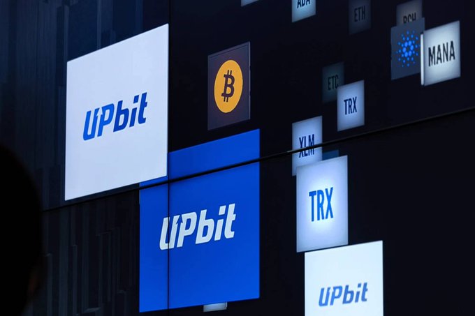 A promotional display for Upbit, South Korea's largest cryptocurrency exchange, features illuminated logos alongside tickers for various coins including BTC, ETH, XRP, TRX, XLM, ADA, and MANA, highlighting the exchange's broad crypto offerings. The prominent branding ties directly to the surge in trading volume—with XRP driving much of the activity, surpassing BTC, USDT, and ETH—reflecting renewed investor interest and the return of the Kimchi Premium on Upbit's platform.