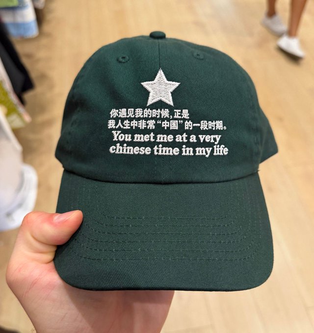 A dark green baseball cap with a white star and embroidered text in Chinese and English reading 'You met me at a very chinese time in my life'.
