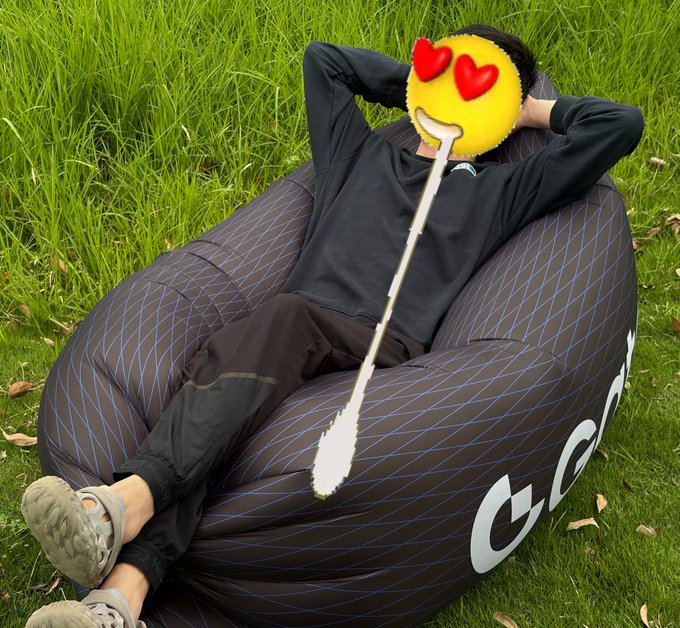 A person relaxing on a black and blue patterned bean bag chair on grass, with their face covered by a heart-eyes emoji sticker and a white scribble drawn over the image.