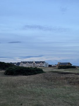 A dusk landscape showing a large building complex in the distance behind grassy fields and shrubs under a cloudy blue sky.
