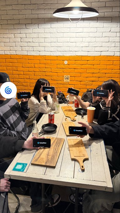 A group of people seated around a long wooden table in a restaurant, all holding up phones displaying a 'TurkeyTether' splash screen.