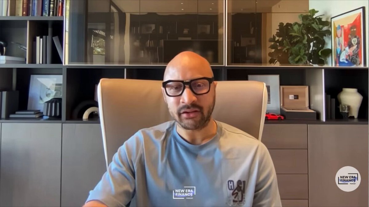 The image shows a man sitting at a desk with glasses on his face, wearing a blue shirt that says "Centralized Companies Have Failed At Protecting Us User Privacy". The background of the image features bookshelves filled with various items such as vases and potted plants. There is also an image of a person in front of the bookshelves.