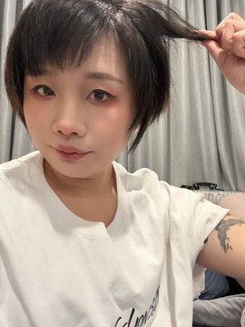 A selfie of a young woman with short dark hair wearing a white t-shirt, pulling a strand of her hair while looking at the camera, with a tattoo visible on her arm.