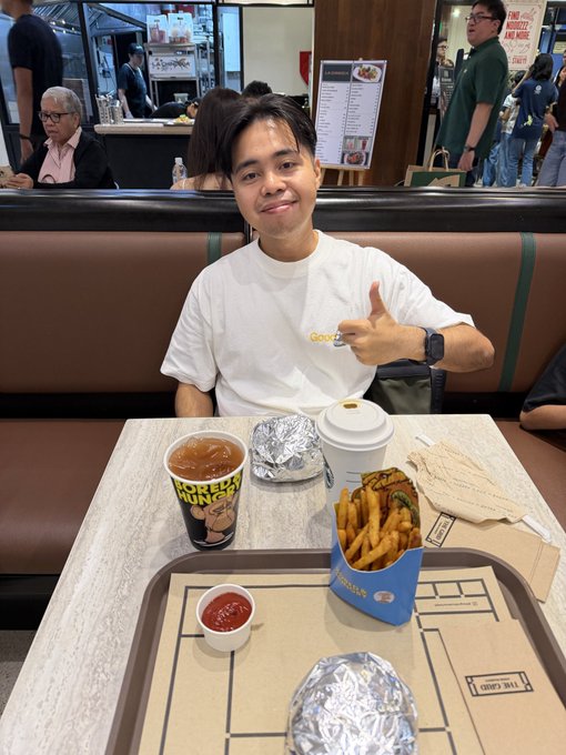 A young man in a white t-shirt sits at a restaurant table giving a thumbs-up, with a meal of a burger, fries, and drinks including a 'Dickhead Dong' branded cup in front of him.