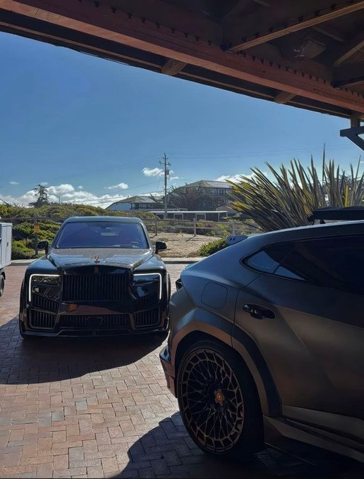 Two luxury SUVs, including a Rolls-Royce, are parked under a wooden pergola on a brick driveway with a scenic landscape and house in the background.