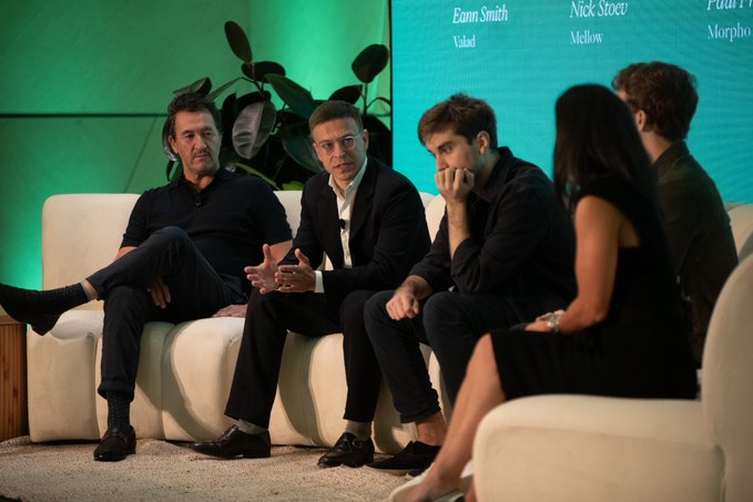 A panel of four men sits on a white couch on stage during a discussion, with visible names including 'Emm Smith' from 'Valad' and 'Nick Steer' from 'Mellow' displayed on a screen behind them.