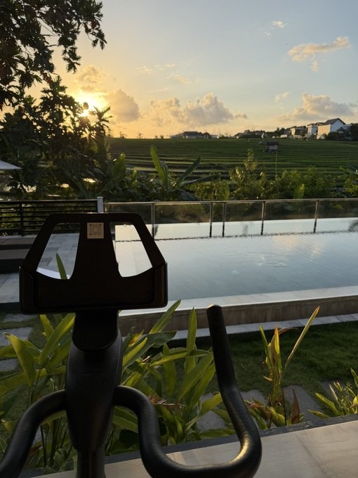 An outdoor viewer's binoculars face a serene infinity pool overlooking rice terraces at sunset, with tropical greenery framing the scene. The image humorously contrasts the peaceful vacation setting with the post's chaotic trading advice, as if the trader has set down their telescope to briefly enjoy the view before ignoring their own wellness tips.