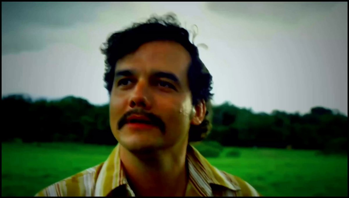 The image shows a man wearing a striped shirt standing in an open field under cloudy skies. The man has a mustache, which adds character to his appearance. He appears to be looking up into the sky with a thoughtful expression.