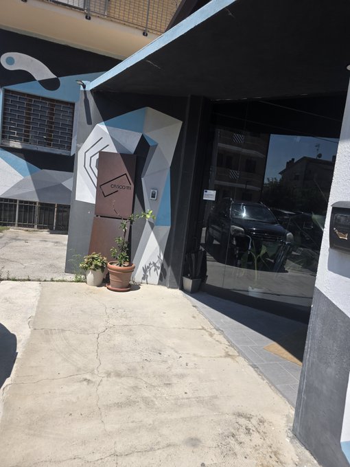 The exterior of a building with black, white, and blue geometric mural art, featuring a glass entrance and a dark car parked nearby.