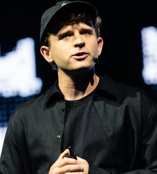 A young man wearing a black cap and shirt is speaking at what appears to be a conference, with blurred text visible in the background. The image captures Jack Mallers, CEO of Strike, articulating his vision for using Bitcoin's decentralized network to liberate individuals from corporate dependency.
