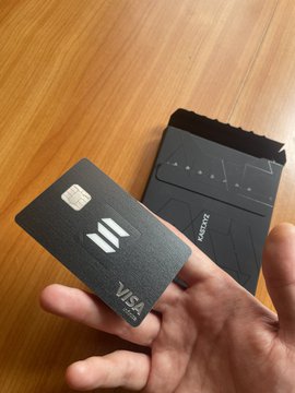 A hand holding a black metal Visa credit card with the Next logo, displayed above its matching black packaging box.