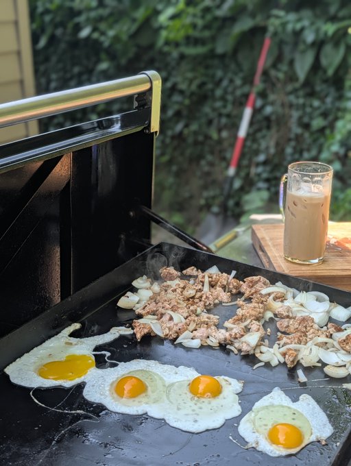 Outdoor cooking scene showing four sunny-side-up eggs and chopped meat with onions cooking on a black griddle, accompanied by an iced coffee drink on a wooden cutting board.