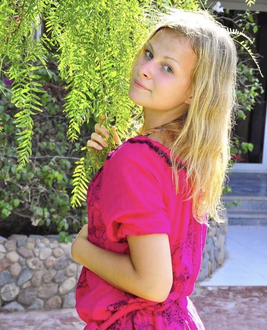 A young blonde girl in a bright pink dress stands outdoors, looking over her shoulder next to a leafy green bush.