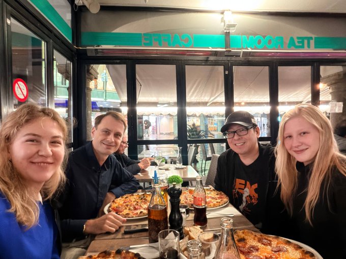 The JAN3 team enjoys pizza and drinks together at a restaurant in Lugano, smiling for a group photo. A no-smoking sign and a reversed “TE ROOM CAFFE” sign are visible in the background.