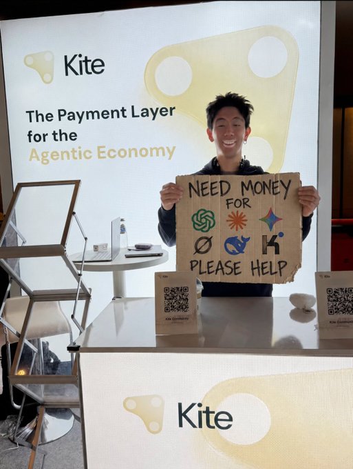 A smiling man holds a handmade sign reading 'NEED MONEY FOR [logos] PLEASE HELP' in front of a booth labeled 'Kite' with text 'The Payment Layer for the Agentic Economy'.