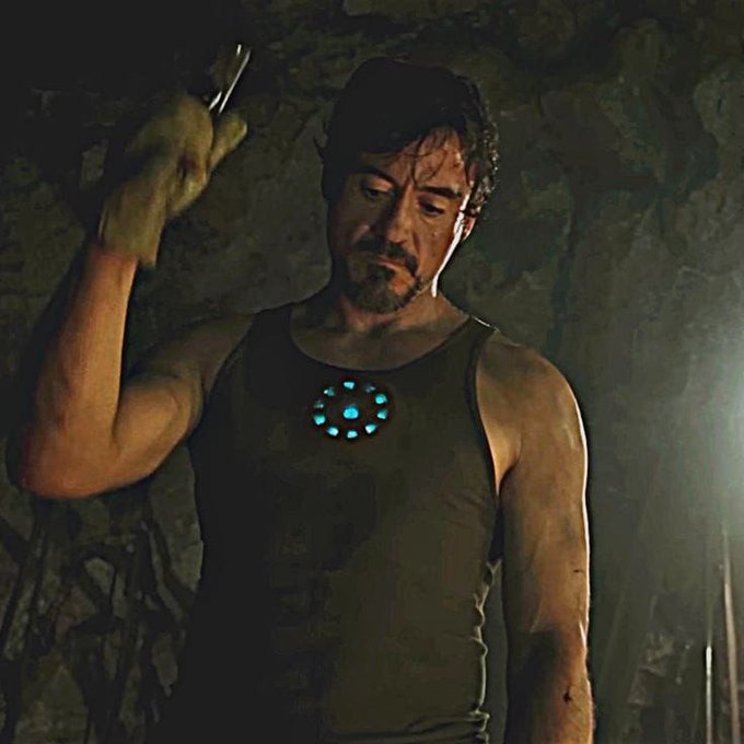 A disheveled Tony Stark stands in his dimly lit cave wearing a tank top with a glowing arc reactor on his chest, holding a wrench as if about to work on something. The image humorously mirrors the caption, portraying him as a stressed developer painstakingly rebuilding his software from scratch at 3am in pursuit of perfection.