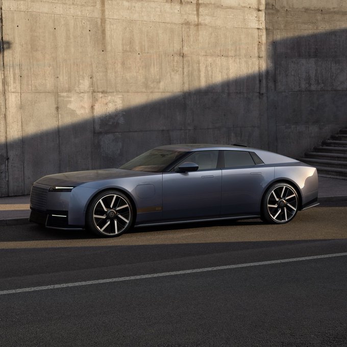 A silver luxury concept sedan is parked on a street in front of a textured concrete wall, illuminated by sunlight casting strong shadows.