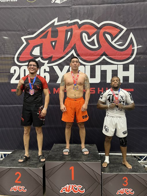 Three competitors stand on the ADCC 2026 Youth Championship podium, with the 1st place winner in orange "USA" shorts proudly displaying his gold medal after his "mission accomplished" victory.