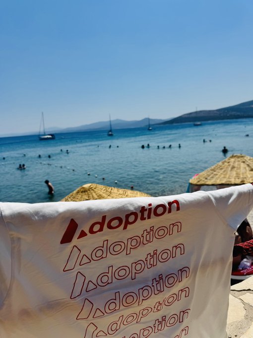 A white t-shirt with the word "dopt" and "doption" repeated in red text is held up in the foreground, with a beach scene featuring turquoise water, swimmers, sailboats, and thatched umbrellas in the background.