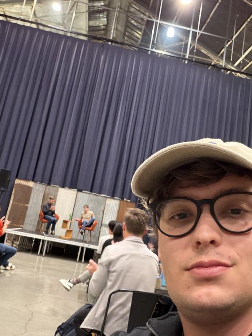 A young man wearing a baseball cap and glasses takes a selfie at what appears to be a panel discussion or convention, with two people seated on chairs on a small stage in the background.