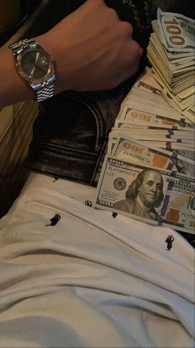 A photograph showing a person's arm wearing a silver Rolex watch resting next to a black bag and multiple stacks of US hundred-dollar bills on a white surface.