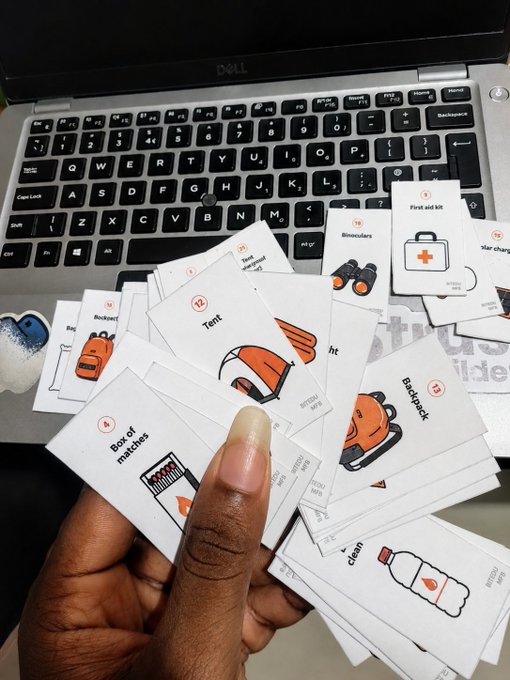 A hand holding a fan of illustrated emergency preparedness cards depicting items like tent, first aid kit, binoculars, rocket, backpack, and box of matches, photographed over a Dell laptop keyboard.