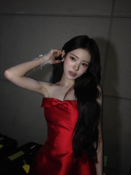 A young woman with long dark hair wearing a red strapless dress, posing indoors against a gray wall while holding up a clear plastic bag.